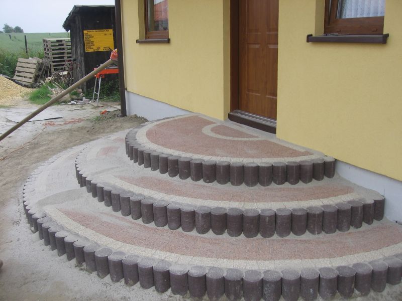 Półokrągłe schody wejściowe do domu w Tychach wykończone kostką w odcieniach beżu i brązu, obrzeża z ciemnych, cylindrycznych elementów. Widoczna żółta elewacja budynku i brązowe okno.